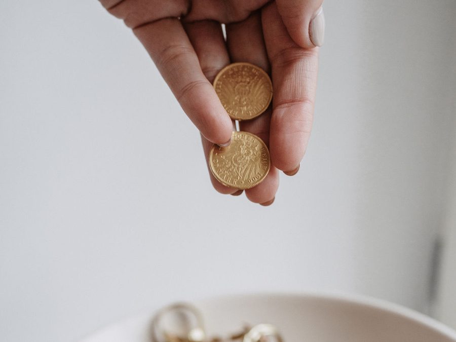 Zwei Finger halten zwei Goldmünzen über einer Schale mit Altgold