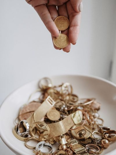 Finger halten eine einzelne Goldmünze über einer Schale mit Altgold