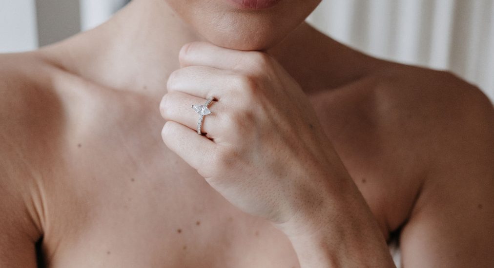 Frau stützt das Kinn auf die Hand, am Ringfinger ein Verlobungsring mit einzelnem Brillanten